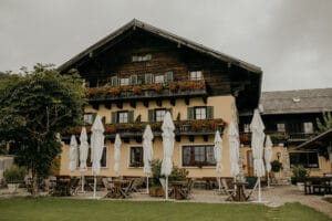 Ein gemütliches, traditionelles Holzhaus mit bunten Blumenkästen unter den Fenstern ist perfekt für eine charmante Hochzeit. Es verfügt über eine Außenterrasse mit geschlossenen weißen Sonnenschirmen und Holztischen und -stühlen, ideal für die Hochzeitsplanung. Der bewölkte Himmel verleiht der Szene einen rustikalen Charme.