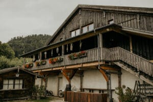 Ein rustikales Holzgebäude mit zwei Stockwerken und einem mit roten Blumen geschmückten Balkon bietet die Bühne für eine bezaubernde Hochzeit. Umgeben von Grün und Bergen verfügt dieser Veranstaltungsort über eine Außentreppe, perfekt für Ihre idyllische Hochzeitsplanung in einer malerischen ländlichen Umgebung.