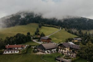 Luftaufnahme einer malerischen Ansammlung von Gebäuden, eingebettet in ein üppiges, grünes Tal, umgeben von nebligen Hügeln und dichten Wäldern. Perfekt für Hochzeitsträume, eine kurvenreiche Straße führt durch die Landschaft und verstärkt die ruhige und malerische Landschaftsszene, ideal für die Hochzeitsplanung.