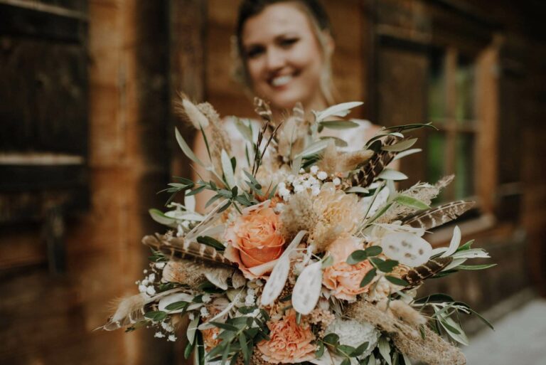 Eine Braut hält einen großen Blumenstrauß mit orangefarbenen Rosen, weißen Blumen, Grünpflanzen und Federn, perfekt für ihre Hochzeit. Im Hintergrund ist eine rustikale Holzkonstruktion zu sehen. Das Gesicht der Braut ist unscharf.