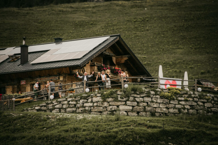 Eine rustikale Holzhütte mit Solarmodulen steht auf einem grasbewachsenen Hügel und ist perfekt für einen traumhaften Hochzeitsausflug. Mehrere Personen stehen auf der Veranda und ein mit Dekorationen und einem herzförmigen Schild geschmückter Zaun im Vordergrund deutet auf romantische Hochzeitsplanung hin.