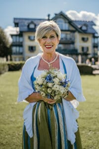 Eine Frau in traditioneller Kleidung steht lächelnd da und hält einen Strauß mit blauen und weißen Blumen in der Hand, der die Essenz einer Hochzeit verkörpert. Im Hintergrund ist ein großes Haus vor einem klaren blauen Himmel zu sehen, eine perfekte Kulisse für jeden Hochzeitsplanungstraum.