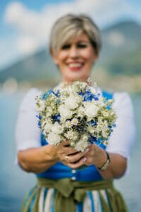 Eine Frau in einem blau-weißen Kleid hält einen Strauß weißer und blauer Blumen und lächelt in die Kamera. Mit einem ruhigen See und verschwommenen Bergen im Hintergrund ist klar, dass ihr Hochzeitstag wunderschön verläuft.