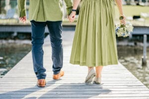 Ein Paar schlendert Händchen haltend an einem Holzsteg entlang und ist in Glückseligkeit versunken. Der Mann trägt blaue Hosen und eine grüne Jacke, während die Frau in einem grünen Kleid einen Blumenstrauß trägt. Diese heitere Szene blickt auf ein ruhiges Gewässer und fängt die Essenz von Liebe und Träumen ein.