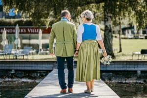 Ein älteres Paar geht Hand in Hand über einen Holzsteg und erinnert an eine zeitlose Hochzeit. Der Mann trägt eine grüne Jacke und ein blaues Hemd, während die Frau, die einen Blumenstrauß hält, elegant in Grün und Blau gekleidet ist. Bäume und Stühle im Hintergrund lassen auf eine intime Feier schließen.