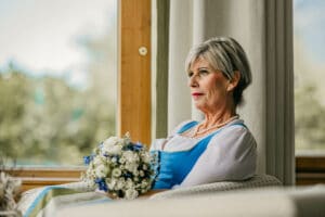 Eine ältere Frau mit kurzen grauen Haaren sitzt in einem Korbstuhl am Fenster. Sie trägt ein traditionelles blau-weißes Kleid mit einer Perlenkette, das an ihren Hochzeitstag erinnert, und hält einen Blumenstrauß in der Hand. Sie sieht nachdenklich aus. Im Hintergrund ist verschwommenes Grün zu sehen.