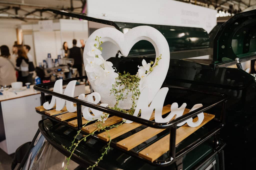 Ein dekorativer Hochzeitsaufsteller auf einem Oldtimer zeigt ein herzförmiges Ornament mit weißen Blumen und grünem Blattwerk. Darunter bilden Holzbuchstaben „Mr & Mrs“ und verkörpern die Essenz eleganter Hochzeitsplanung. Die Kulisse scheint drinnen zu sein, mit verschwommenen Veranstaltungsteilnehmern im Hintergrund.