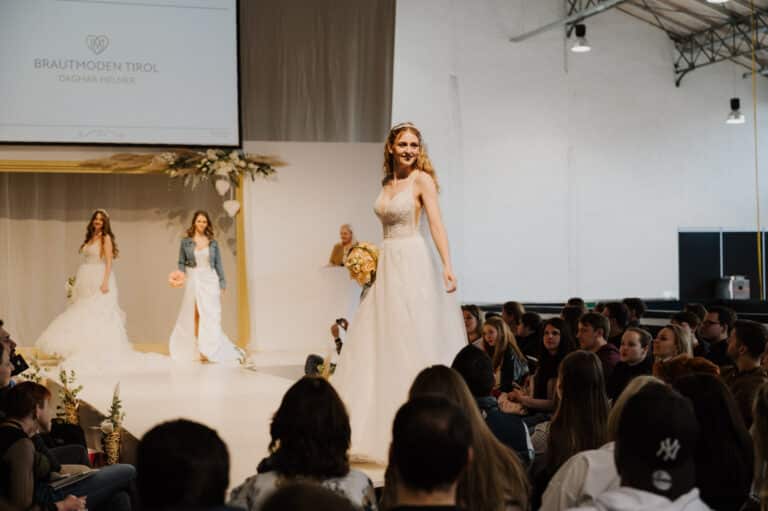 Eine Braut in einem weißen Kleid läuft selbstbewusst über den Laufsteg einer Modenschau, lächelt und hält einen Blumenstrauß in der Hand – die Verkörperung der Träume aller, die ihre Hochzeit planen. Ein weiteres Model folgt ihr, während das Publikum aufmerksam zusieht. Auf einem Bildschirm wird „Brautmoden Tirol Dagmar Nemeier“ gezeigt, was die Essenz des Heiratens einfängt.