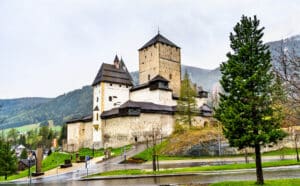 Eine mittelalterliche Burg mit mehreren Türmen steht auf einem felsigen Hügel, umgeben von immergrünen Bäumen, und bietet eine märchenhafte Kulisse, die perfekt für eine Traumhochzeit ist. Der bewölkte Himmel und die nasse Straße tragen zur romantischen Atmosphäre bei, während die Berge diese zauberhafte Szene majestätisch einrahmen.