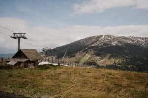 Eine rustikale Holzhütte steht neben einem Skilift auf einem grasbewachsenen Berghang und bietet mit schneebedeckten Gipfeln und grünen Wäldern im Hintergrund unter einem teilweise bewölkten Himmel die perfekte Kulisse für Ihre Hochzeit.