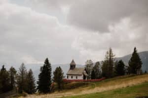 Eine kleine weiße Kapelle mit Holzdach und Kirchturm steht auf einem Hügel, umgeben von hohen immergrünen Bäumen. Ein wolkiger Himmel und Berge in der Ferne bilden die Kulisse, rote Blumen säumen den Weg – perfekt für alle, die ihre Traumhochzeit in dieser malerischen Umgebung planen.