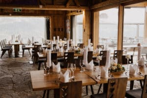 Rustikales Restaurant-Setup mit Holztischen und -stühlen, perfekt für Hochzeitsplanung. Herzförmige Stuhllehnen und große Fenster geben den Blick auf die Berge frei und bieten eine einladende Atmosphäre, ideal für diejenigen, die davon träumen, in einer gemütlichen Umgebung zu heiraten.