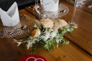 Ein Blumenarrangement aus hellen Pfirsichrosen mit Grünpflanzen und weißen Akzenten schmückt einen Holztisch, perfekt für eine intime Hochzeit. Daneben stehen Weingläser, gefaltete Servietten und ein roter runder Gegenstand mit ineinander verschlungenen weißen Ringen, die der Hochzeitskulisse Eleganz verleihen.