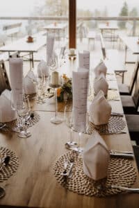 Ein wunderschön gedeckter Esstisch für eine Hochzeit, mit gerollten Menüs, ordentlich gefalteten Servietten, Weingläsern und Silberbesteck. Der Tisch ist mit einem Blumenschmuck geschmückt, während große Fenster einen Blick auf den bezaubernden Sitzbereich im Freien bieten.