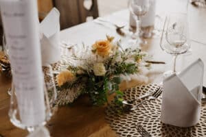 Ein wunderschön gedeckter Esstisch für eine Hochzeit verfügt über zarte florale Tischdekorationen mit pfirsichfarbenen und weißen Rosen, elegante Weingläser und ordentlich gefaltete weiße Servietten auf aufwendig gestalteten Tischsets – perfekt für jedes Paar, das schon mitten in der Hochzeitsplanung steckt.