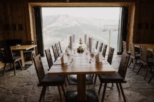 Ein rustikaler Speisesaal, der sich perfekt für eine Hochzeit eignet, bietet Aussicht auf eine verschneite Berglandschaft. Holzstühle stehen um Tische herum, die mit gefalteten Servietten, Gläsern und Speisekarten geschmückt sind. Lichterketten rahmen das große Fenster ein und bieten eine malerische Aussicht, die ideal für Paare ist, die heiraten möchten.