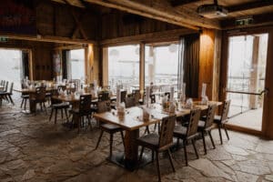 Ein rustikales Restaurant-Interieur, perfekt für eine Hochzeit, mit Steinboden und Holzwänden. Mehrere Holztische sind elegant mit gefalteten Servietten und Gläsern gedeckt. Große Fenster bieten einen Blick nach draußen und lassen den Raum mit natürlichem Licht durchfluten – ein idealer Rahmen für die Hochzeitsplanung.