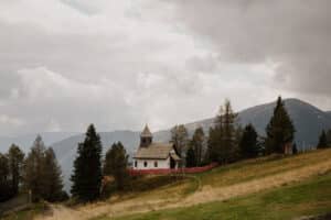 Eine kleine Holzkirche, die sich perfekt für eine Hochzeit eignet, steht umgeben von immergrünen Bäumen auf einem grasbewachsenen Hügel unter einem bewölkten Himmel. Berge bilden die Kulisse, Reihen roter Blumen säumen den Weg zu diesem idyllischen Ort für diejenigen, die ihre Traumhochzeit planen.