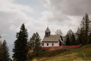 Eine kleine weiße Kirche mit Holzdach steht auf einem grasbewachsenen Hügel, umgeben von Kiefern – perfekt für alle, die von einer intimen Hochzeit träumen. Rote Blumen säumen einen Zaun um die Kirche und schaffen eine zauberhafte Kulisse. Der bewölkte Himmel und die Berge im Hintergrund tragen zur romantischen Atmosphäre bei.