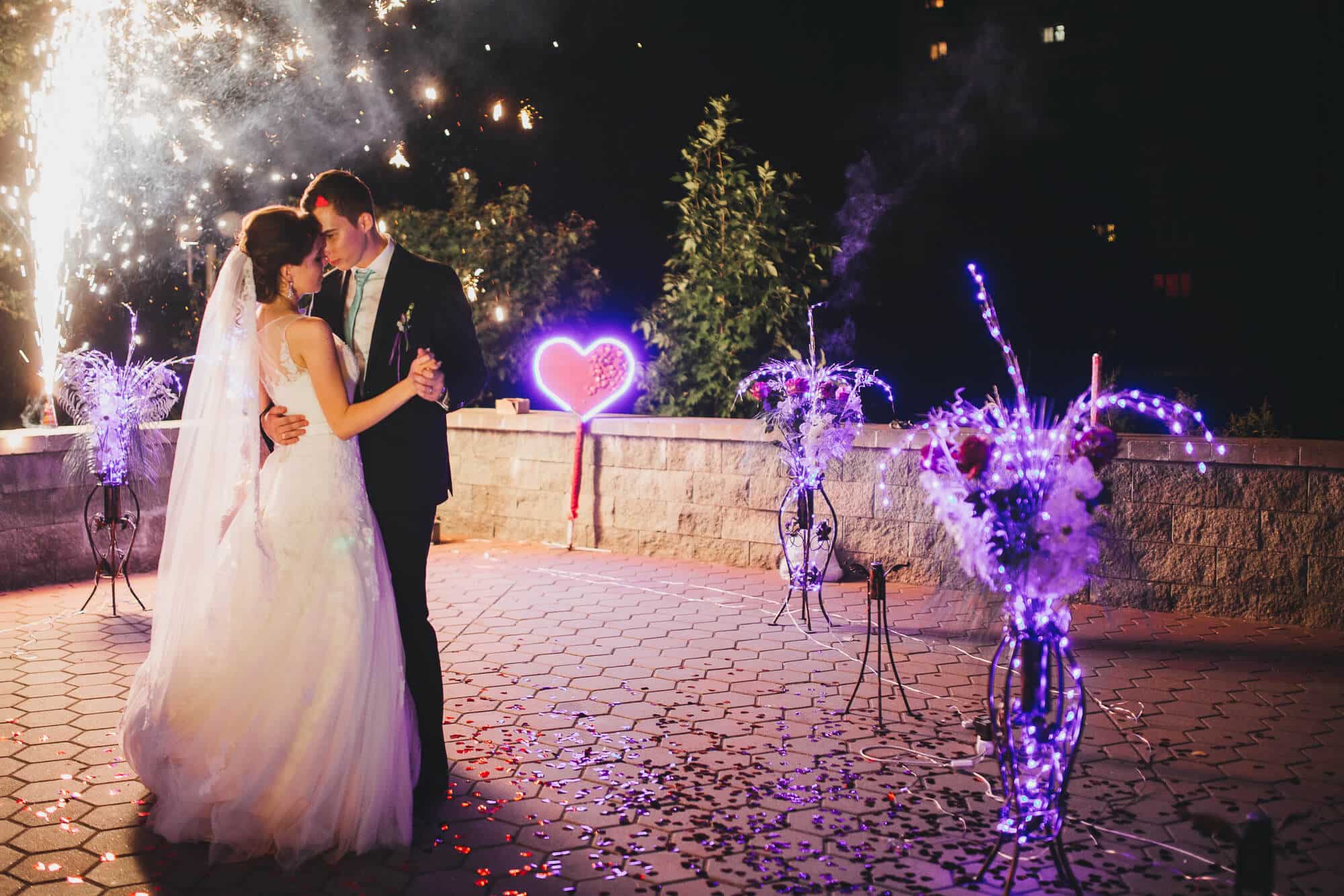 Braut und Bräutigam tanzen abends im Freien ihren ersten Tanz. Sie sind umgeben von Feuerwerk, Blumenarrangements mit violetten Lichtern und einer leuchtenden Herzdekoration. Die Atmosphäre ist festlich und romantisch.