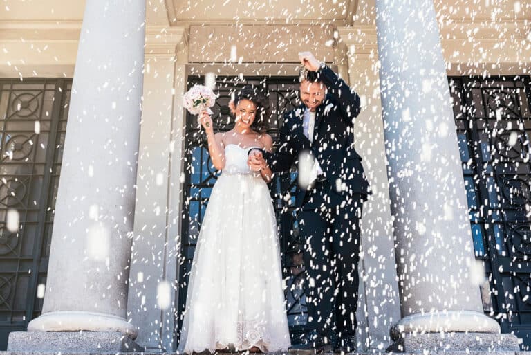 Ein fröhliches Paar im Hochzeitskleid steht auf Stufen zwischen großen Säulen und lächelt inmitten eines Konfettiregens. Die Braut hält einen Blumenstrauß, während der Bräutigam beim festlichen Reiswerfen seinen Arm zum Jubeln streckt.