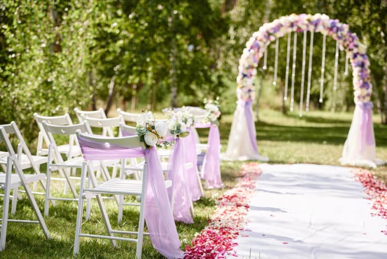 Eine Hochzeit im Freien besteht aus einer wunderschönen Sitzordnung mit weißen Stühlen, die mit violetten Schärpen geschmückt und in Reihen angeordnet sind. Ein weißer Läufer, der mit rosa Blütenblättern gesäumt ist, führt zu einem Blumenbogen, der mit hängenden Kristallen geschmückt ist. Grün umgibt die sonnige Szene.