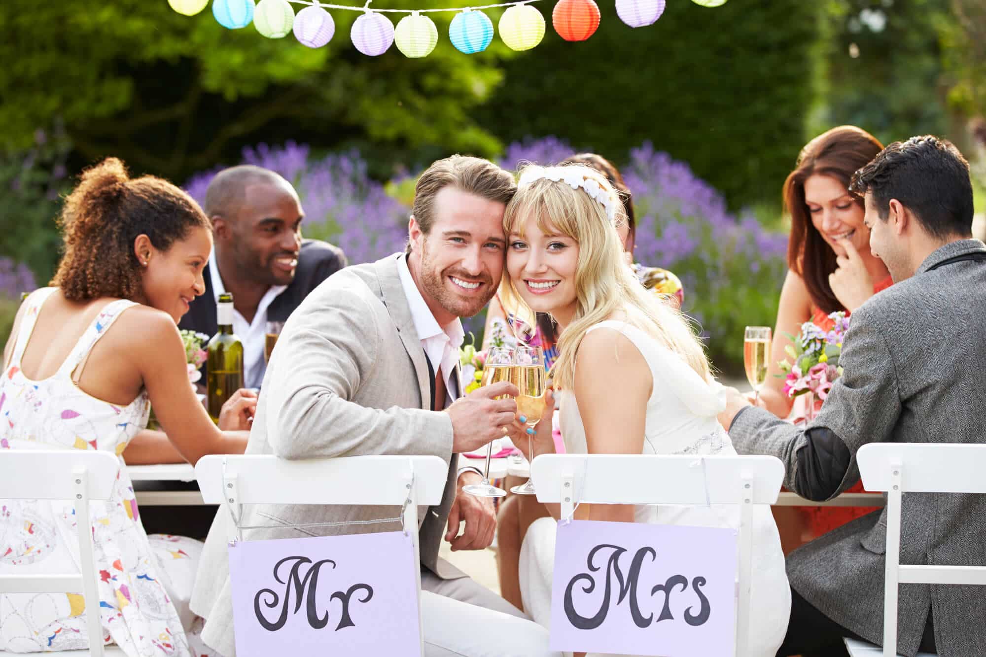 Ein lächelndes Paar in Hochzeitskleidung sitzt an einem dekorierten Empfangstisch im Freien, der an einen fröhlichen Hochzeitszug erinnert. Sie halten Champagnergläser in den Händen und sind von Freunden umgeben. Auf den Stühlen stehen Schilder mit „Mr.“ und „Mrs.“ und über ihnen wiegen sich bunte Laternen vor der Kulisse eines üppigen Gartens.