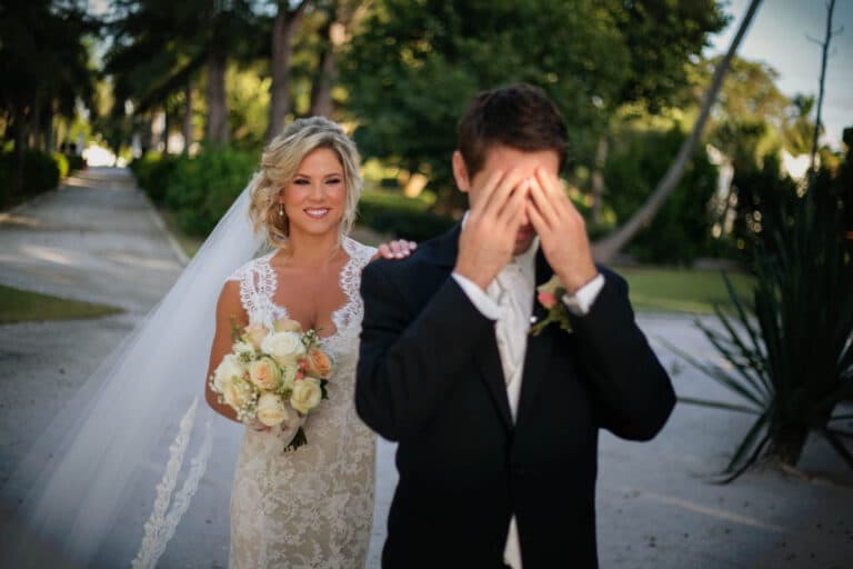 Eine Braut in einem weißen Spitzenkleid und Schleier hält einen Strauß Rosen in der Hand und strahlt bei ihrem ersten Blick, während sie hinter dem Bräutigam steht, der verspielt sein Gesicht mit den Händen bedeckt. Sie stehen draußen auf einem malerischen Weg, der von Bäumen gesäumt ist.