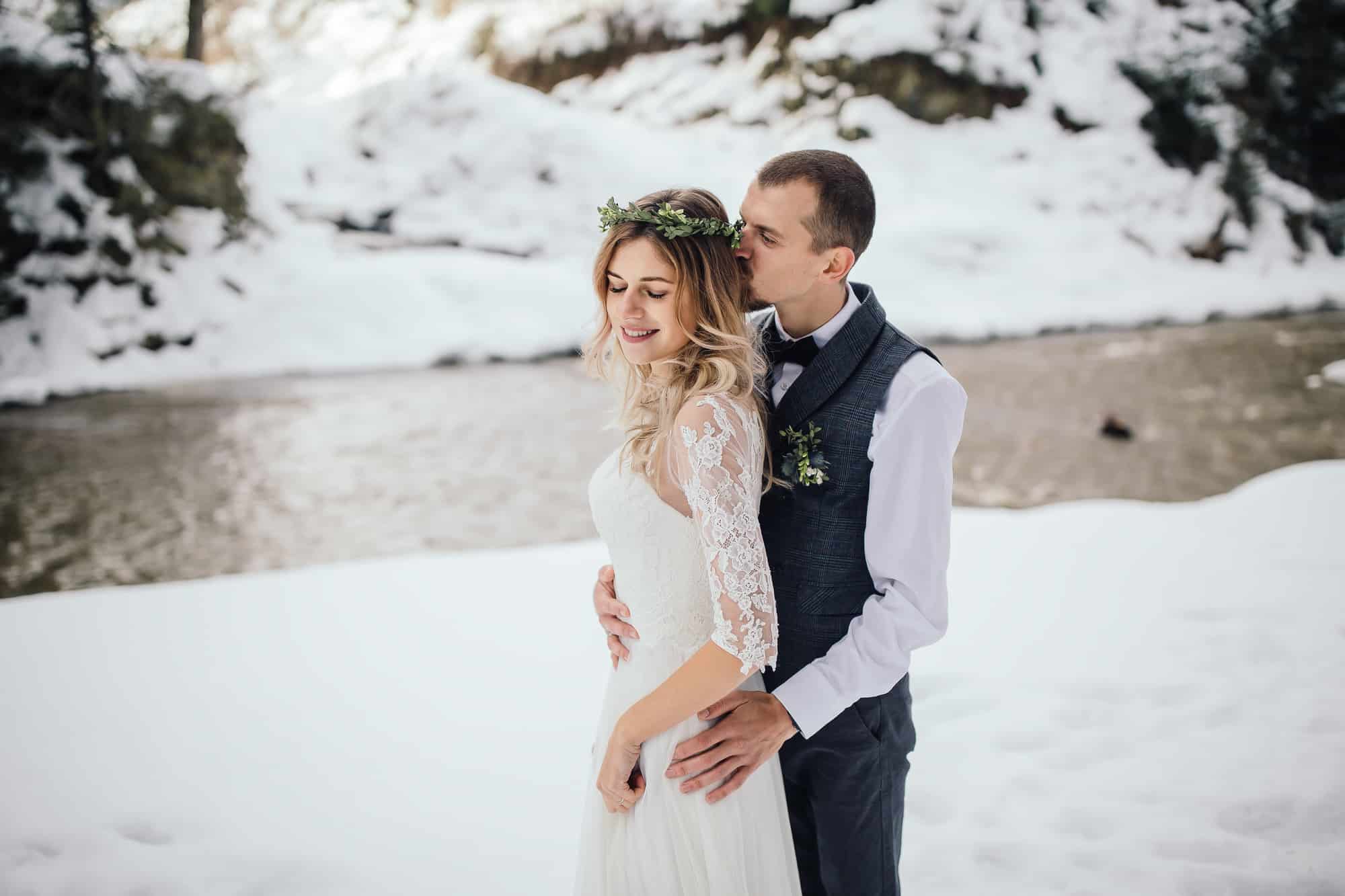 Braut und Bräutigam umarmen sich an einem verschneiten Flussufer, zusammengeführt durch ihre sorgfältige Hochzeitsplanung. Die Braut in einem Kleid mit Spitzenärmeln und einem Blumenkopfschmuck lächelt, während der Bräutigam, der eine Weste und eine Ansteckblume trägt, ihr vor einer Kulisse schneebedeckter Bäume sanft einen Kuss auf den Kopf gibt.