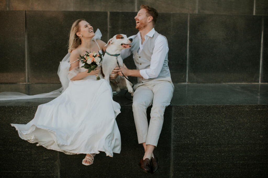 Ein fröhliches Paar sitzt auf einem Steinsims. Die Frau im Brautkleid mit einem Blumenstrauß in der Hand lacht mit dem Mann im hellen Anzug. Zwischen ihnen sitzt ein glücklicher Hund. Der Hintergrund ist eine graue, strukturierte Wand.