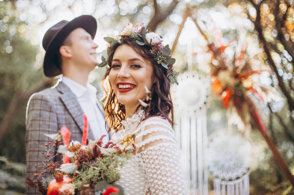 Eine lächelnde Frau in einem Spitzenkleid, das die Brautkleid-Trends 2025 widerspiegelt, und einer Blumenkrone hält einen Blumenstrauß in der Hand. Sie steht in einer Außenkulisse mit verschwommenen Bäumen und Traumfängern. Im Hintergrund steht ein Mann in kariertem Anzug und Hut, der nach oben blickt.