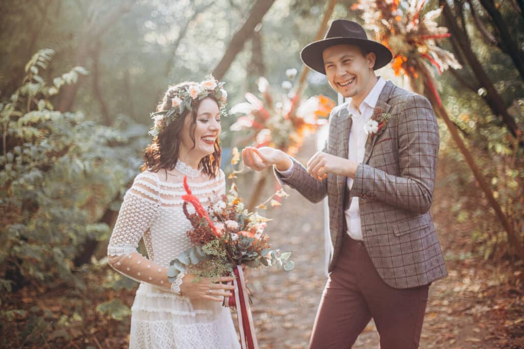 Ein Paar, in Hochzeitskleidung gekleidet und Brautkleid-Trends 2025 umarmend, steht auf einem Waldweg. Die Frau hält einen Blumenstrauß und trägt einen Blumenkranz, während ihr Tüllkleid das Sonnenlicht einfängt, das durch die Bäume fällt. Der Mann, der einen Hut trägt, interagiert spielerisch mit ihr inmitten der Umarmung der Natur.