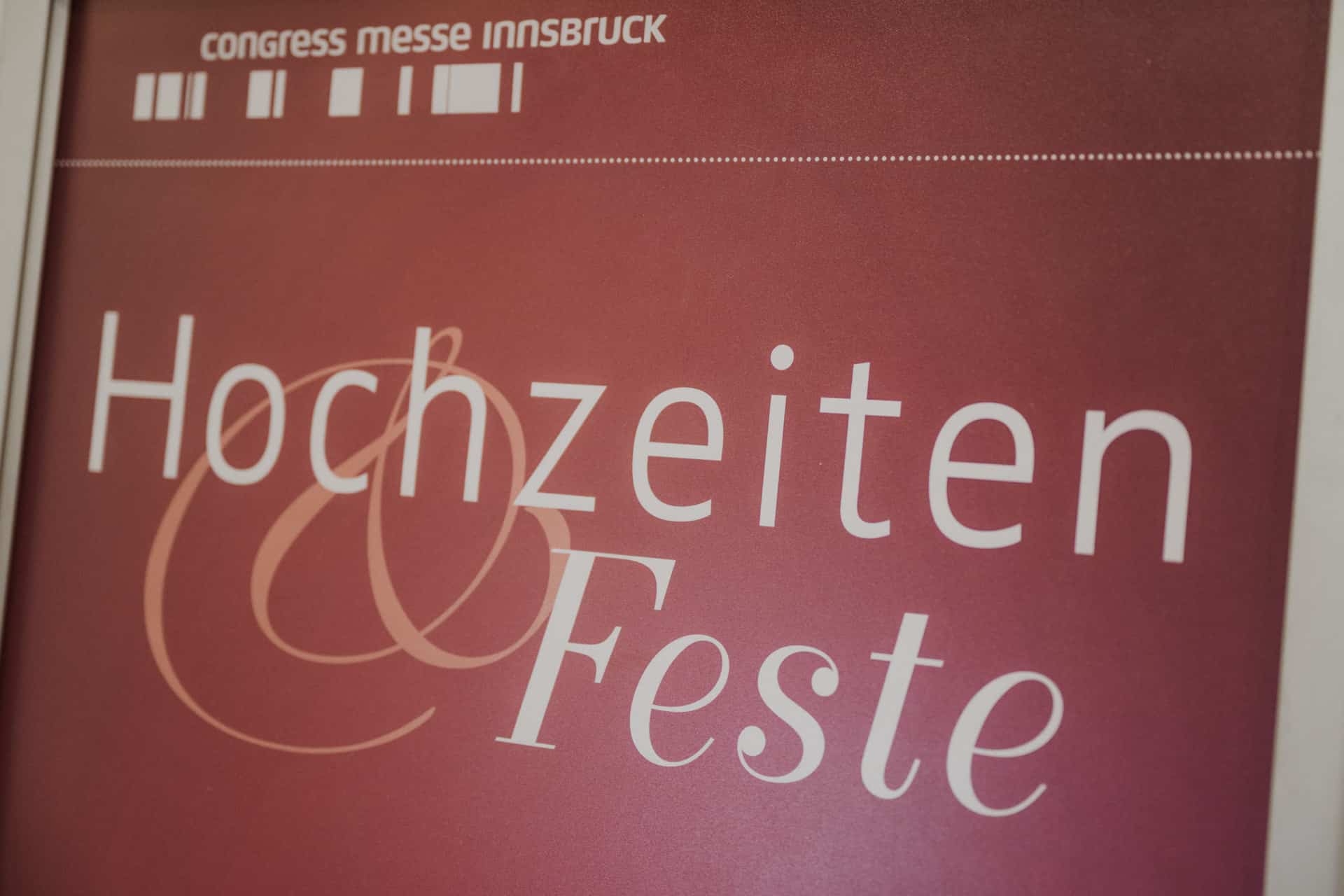 Auf einem burgunderfarbenen Schild steht elegant in fetten weißen Buchstaben „Hochzeiten & Feste“, mit „Congress Messe Innsbruck“ an der Spitze und einem dekorativen Schnörkel, der das Design vervollständigt.