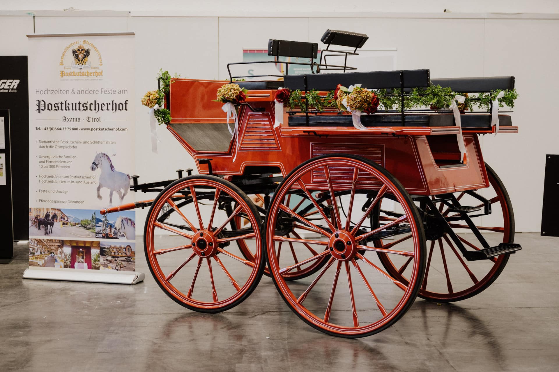 Im Innenbereich wird eine rote, historische Pferdekutsche mit schwarzen Sitzen und Blumenschmuck ausgestellt. Daneben sorgt ein großes Banner mit Veranstaltungsdetails und einem Pferdebild für Charme und schafft eine perfekte Kulisse für Hochzeiten & Feste auf dem Messegelände.