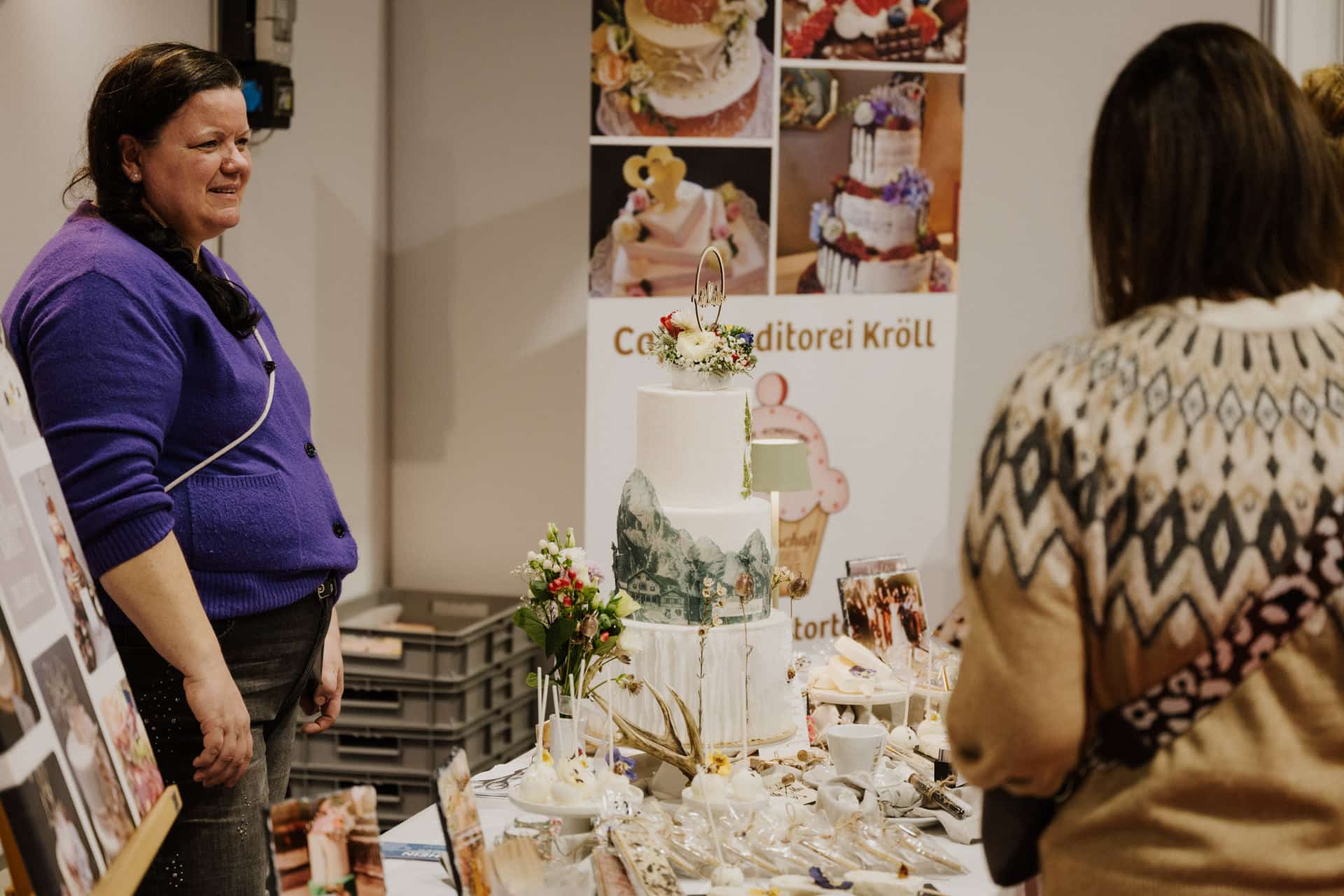 Eine Frau in einem lila Pullover steht neben einem Ausstellungstisch mit einer mehrstöckigen Torte, geschmückt mit Blumen, perfekt für Hochzeiten. Zwei Personen in gemusterten Pullovern bewundern die festliche Szene. Im Hintergrund sind Poster von Torten und anderen festlichen Leckereien zu sehen.