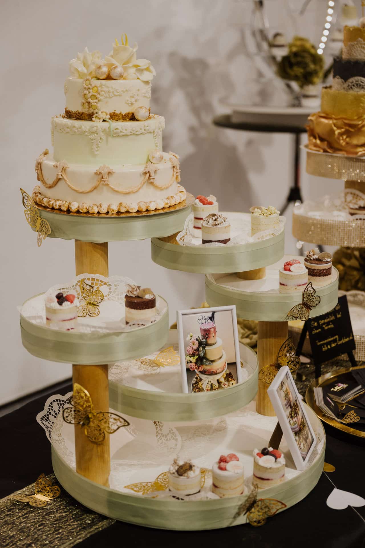 Ein mehrstöckiger Dessertständer hält eine elegante weiße Torte, die mit Blumen geschmückt ist, perfekt für Hochzeiten. Um die Torte herum sind verschiedene kleine Cupcakes mit dekorativen Schmetterlingen und Bilderrahmen angeordnet, was eine anspruchsvolle und künstlerische Präsentation ergibt, die sich ideal für festliche Feiern eignet.