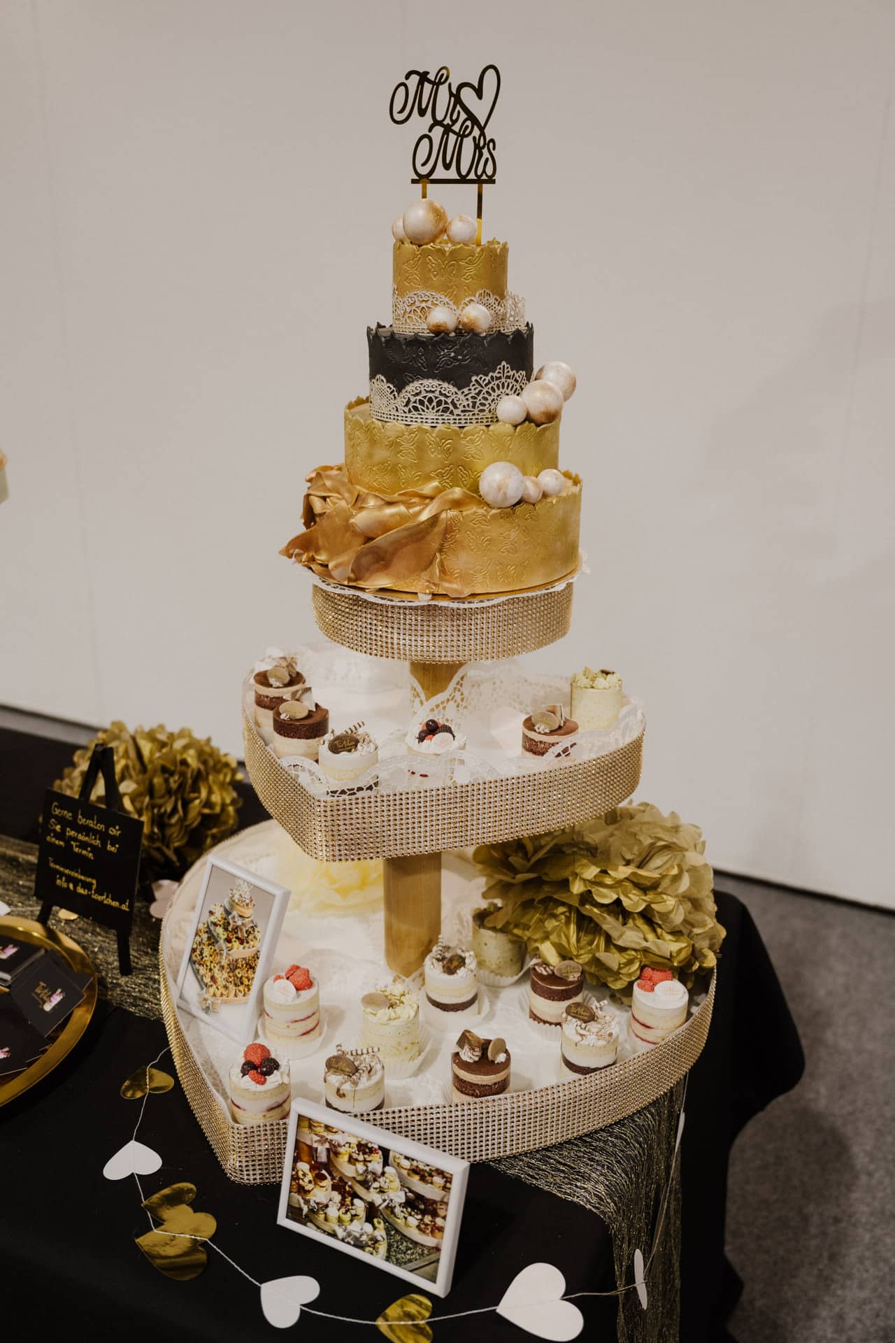 Eine dreistöckige goldene Hochzeitstorte mit dekorativen Blumen und einem „Mr & Mrs“-Aufsatz steht elegant inmitten eines Tisches aus Cupcakes, Fotos und herzförmigen Girlanden und schafft mit ihren charmanten goldenen Akzenten den perfekten Rahmen für Hochzeiten & Feste.