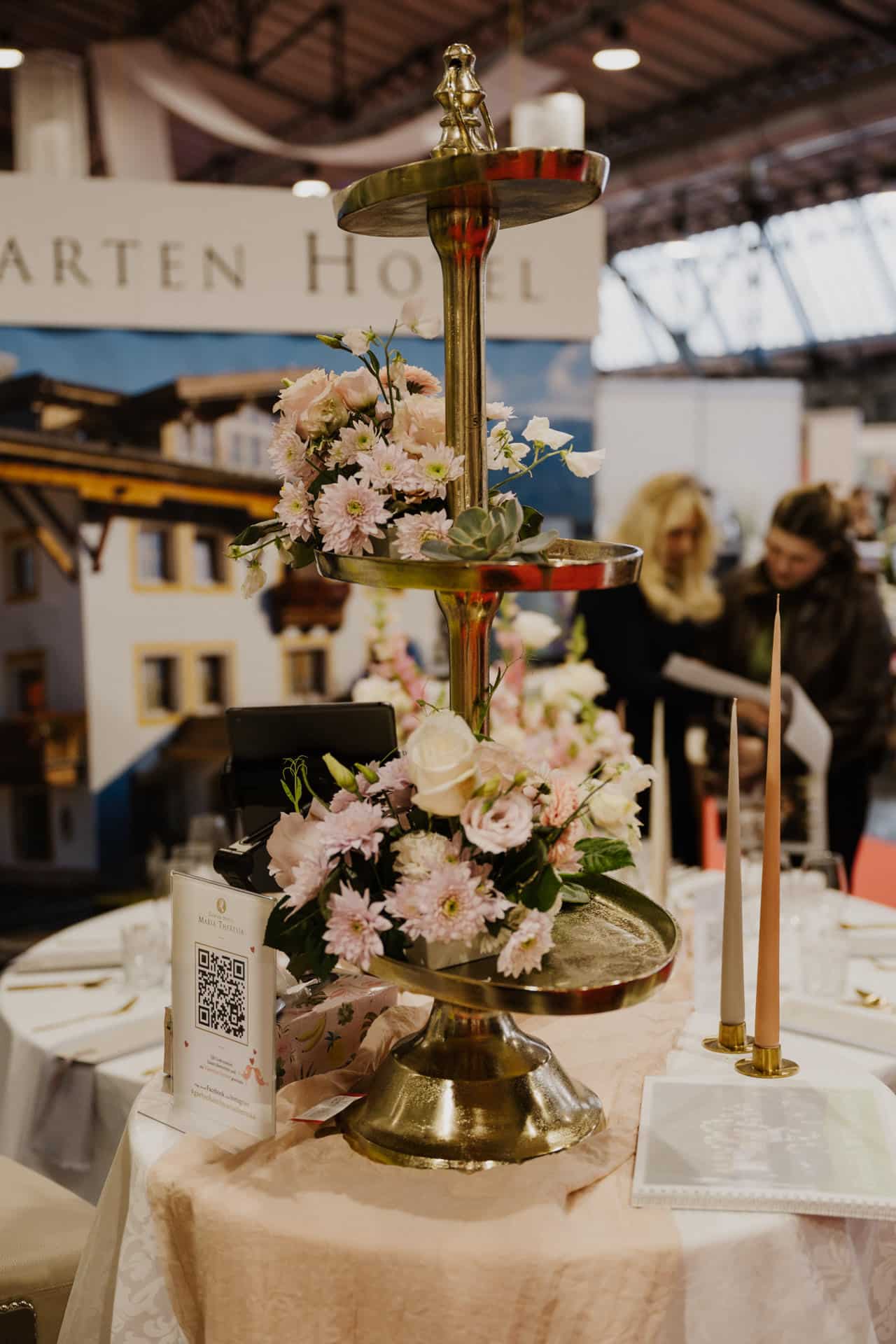 Ein dekorativer, dreistufiger goldener Ständer mit rosa und weißen Blumen ziert einen mit einer rosa Tischdecke geschmückten Tisch, perfekt für Hochzeiten und Feste. In der Nähe flackern zwei große Kerzen neben einem QR-Code und einer Broschüre. Im unscharfen Hintergrund mischen sich Menschen in der Nähe eines Schilds mit teilweisem Text.