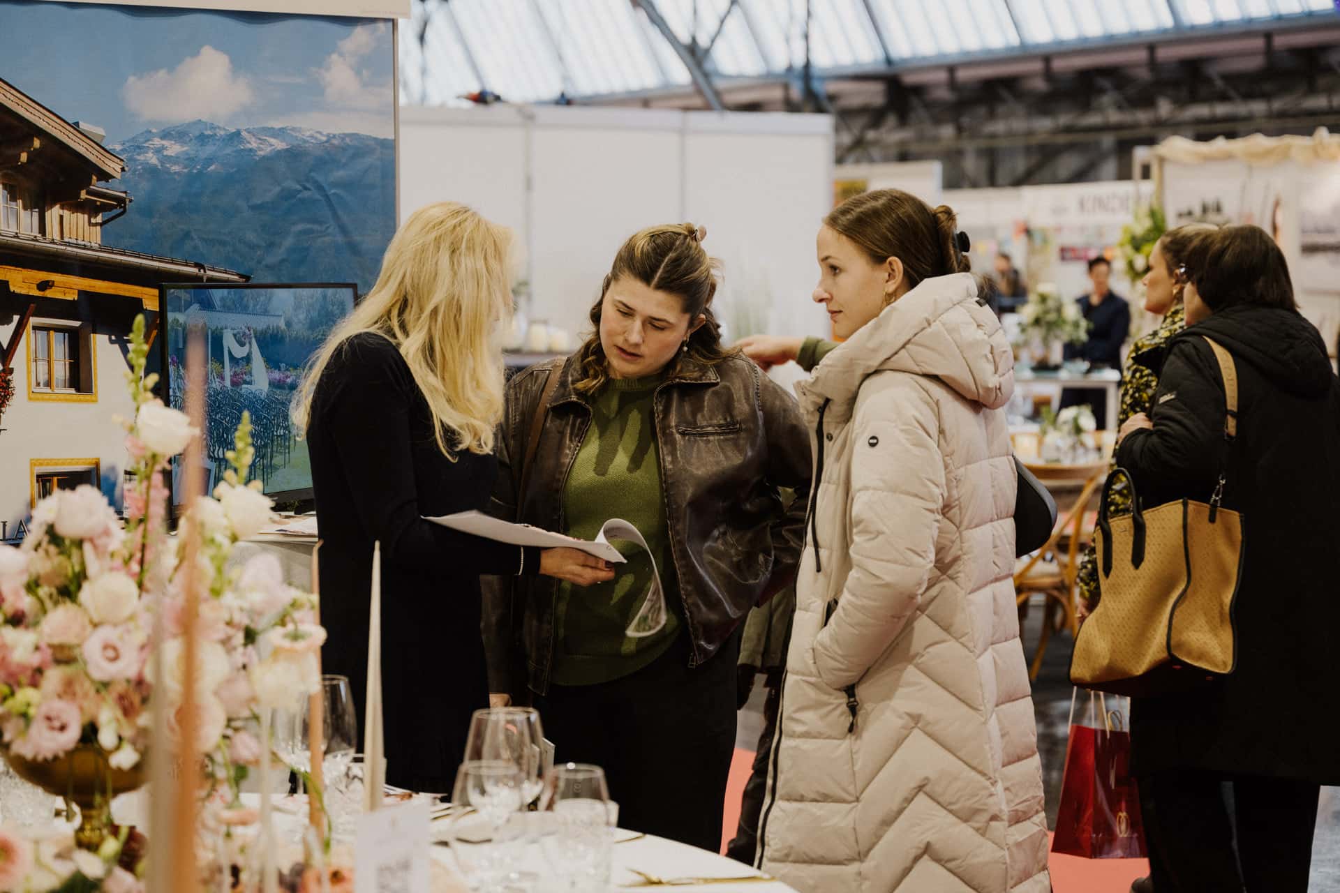 Eine Gruppe dreier Frauen unterhält sich mit einer blonden Frau, die einen Ordner in der Hand hält. Sie stehen an einem mit Blumenschmuck und Broschüren geschmückten Stand von Hochzeiten & Feste in einem geräumigen Innenraum, möglicherweise auf einer Tagung oder Ausstellung.