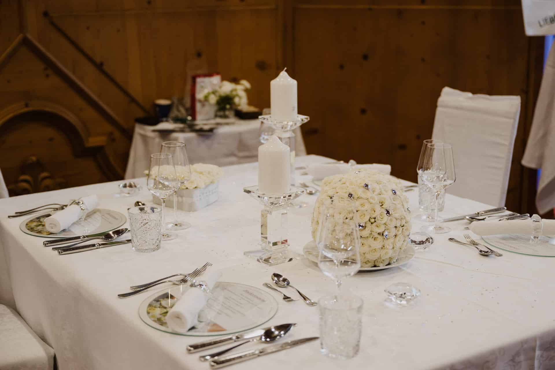 Elegantes Esstischset für Hochzeiten oder festliche Anlässe mit weißer Tischdecke, Kerzen, Kristallgläsern und Silberbesteck. Ein runder Blumenschmuck wird durch gerollte Servietten auf glänzenden Glastellern ergänzt und sorgt für ein gehobenes Ambiente.