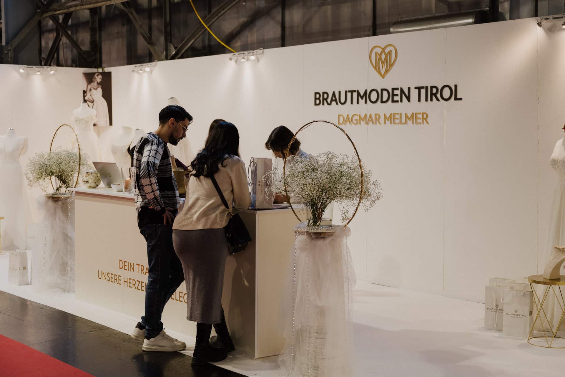 Ein Paar bespricht seine Traumhochzeit mit einer Vertreterin am Brautmodenstand von „Brautmoden Tirol, Dagmar Melmer“. Umgeben von eleganten Brautkleidern und Blumenarrangements fängt der Stand den bezaubernden Geist von Hochzeiten & Feste auf dieser geschäftigen Brautmesse ein.