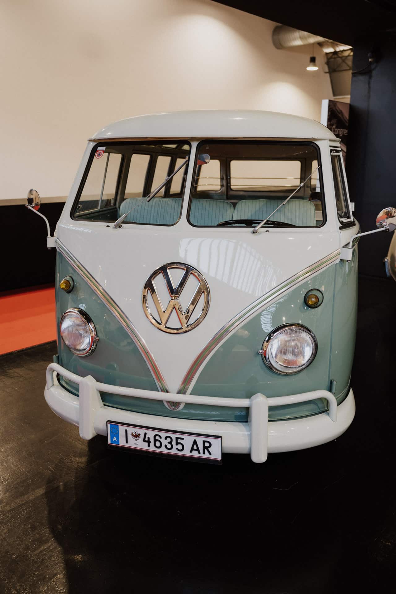 Ein hellblau-weißer Volkswagen-Campingbus im Vintage-Stil mit großem VW-Logo auf der Vorderseite steht für Hochzeiten und Feste bereit. Er verfügt über eine geteilte Windschutzscheibe und ein europäisches Nummernschild und steht drinnen auf einem glänzenden schwarzen Boden.