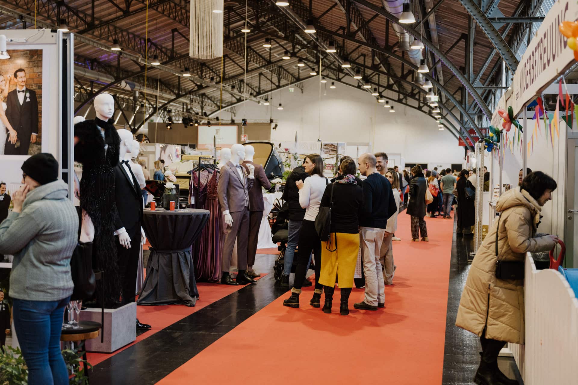 Besucher schlendern durch eine überdachte Ausstellungshalle, in der an Ständen Schaufensterpuppen in eleganter Hochzeitskleidung zu sehen sind. Ein roter Teppich leitet die geschäftige Menge, während Deckenbeleuchtung die festliche Atmosphäre unterstreicht.