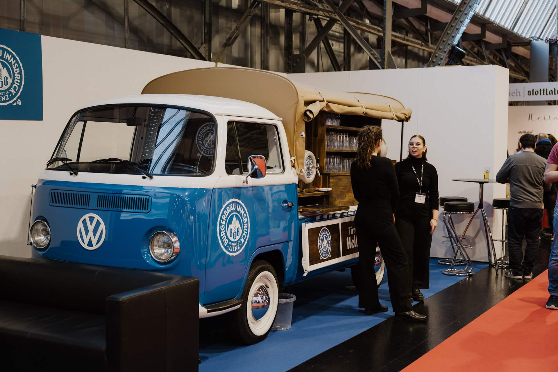 Ein blau-weißer VW-Van im Vintage-Stil, der heute eine schicke mobile Bar mit Theke ist, steht bei einer Veranstaltung im Innenbereich. Zwei schwarz gekleidete Personen unterhalten sich neben dem Van, während sich im Hintergrund andere Gäste unterhalten – perfekt für Hochzeiten und Feste.