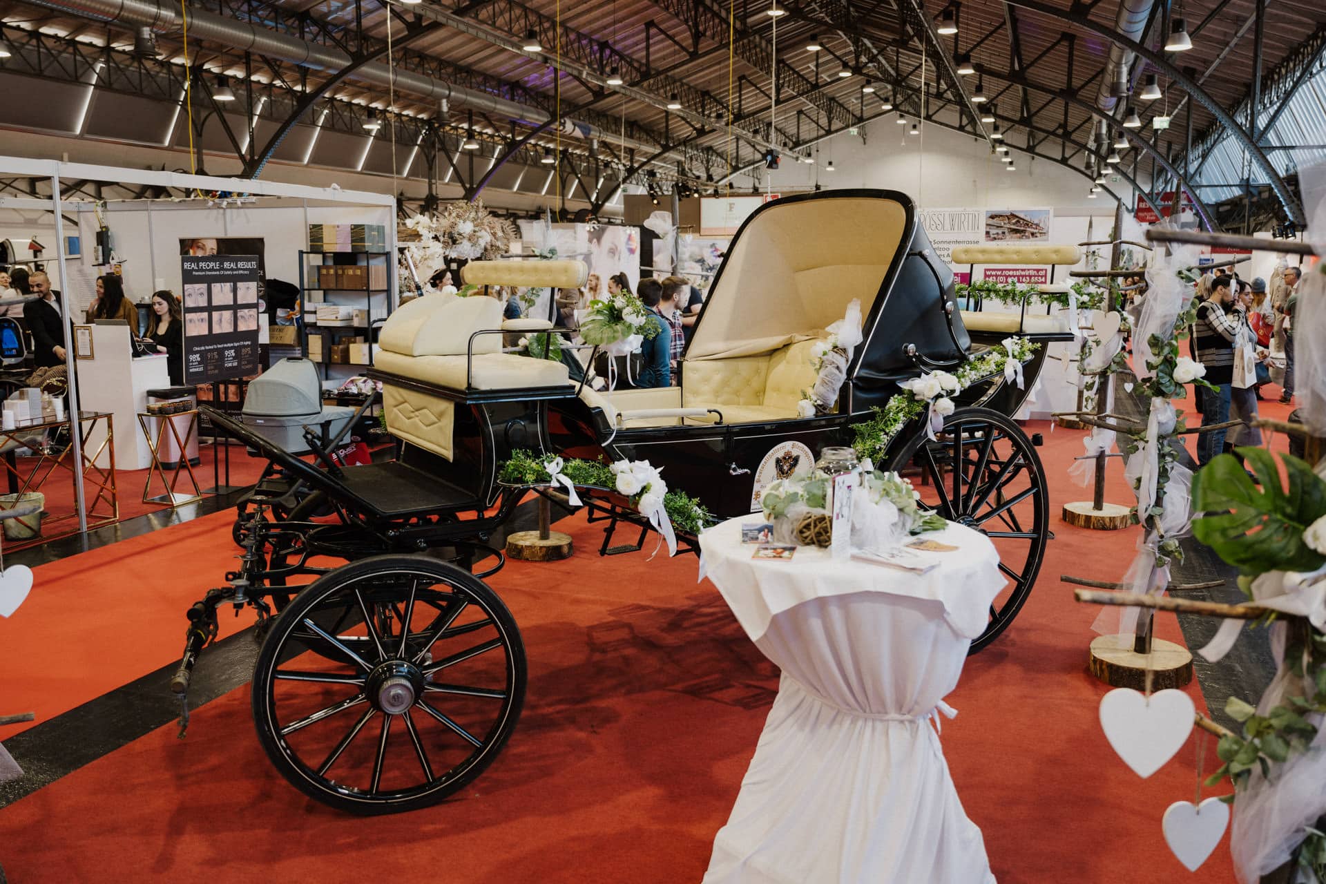 Eine alte Kutsche mit cremefarbener Polsterung wird auf einem roten Teppich in einer Ausstellungshalle präsentiert und spiegelt die Eleganz von Hochzeiten & Feste wider. In der Nähe sorgt ein mit weißem Stoff bedeckter und mit Blumen geschmückter Tisch für Charme, während die Leute im Hintergrund die bezaubernde Atmosphäre genießen.