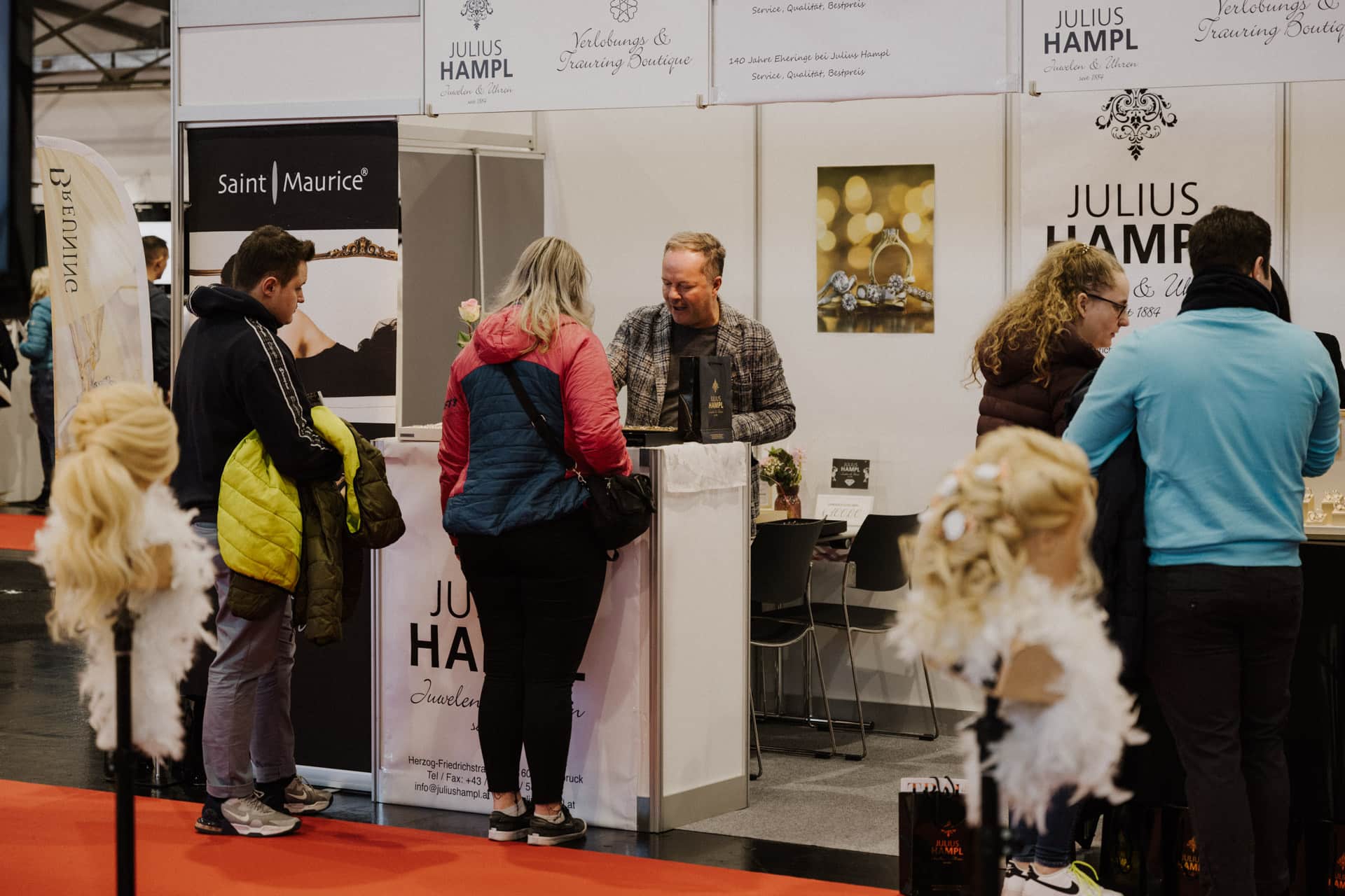 An einem Messestand mit Schildern mit der Aufschrift „Julius Hampl“ und „Saint Maurice“ sprechen Besucher eifrig mit einem Verkäufer. In der Nähe fallen Perücken und Haarteile auf eleganten Ständen ins Auge, die perfekt dazu geeignet sind, Hochzeiten & Feste Flair zu verleihen.