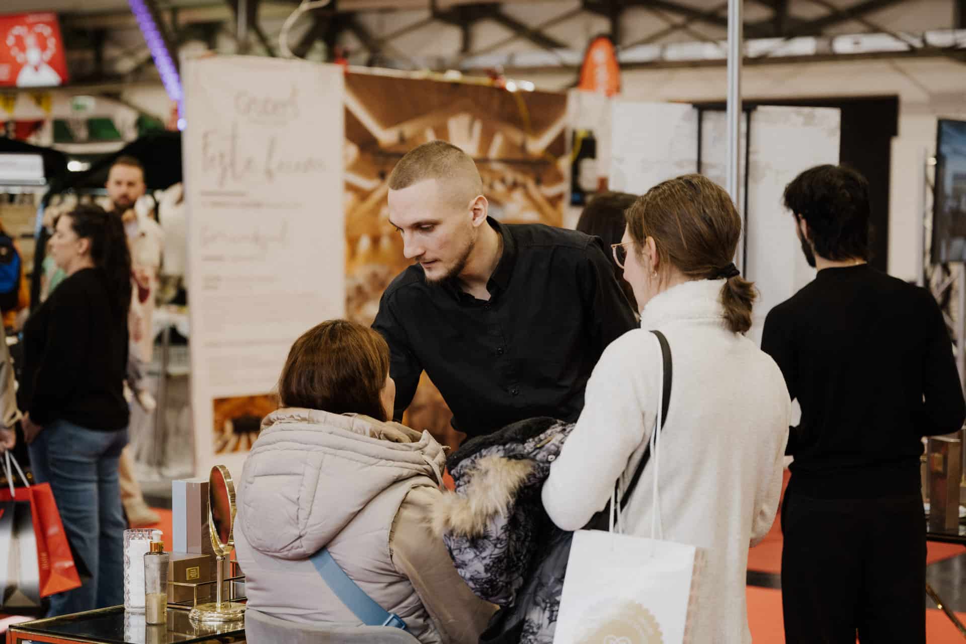 Ein Mann in einem schwarzen Hemd unterhält sich mit zwei Frauen auf einer geschäftigen Messe oder einem Markt, der sich auf „Hochzeiten & Feste“ konzentriert. Die Szene ist lebhaft, die Leute mischen sich und farbenfrohe Plakate, Stände und Banner bieten eine festliche Kulisse für den Anlass.