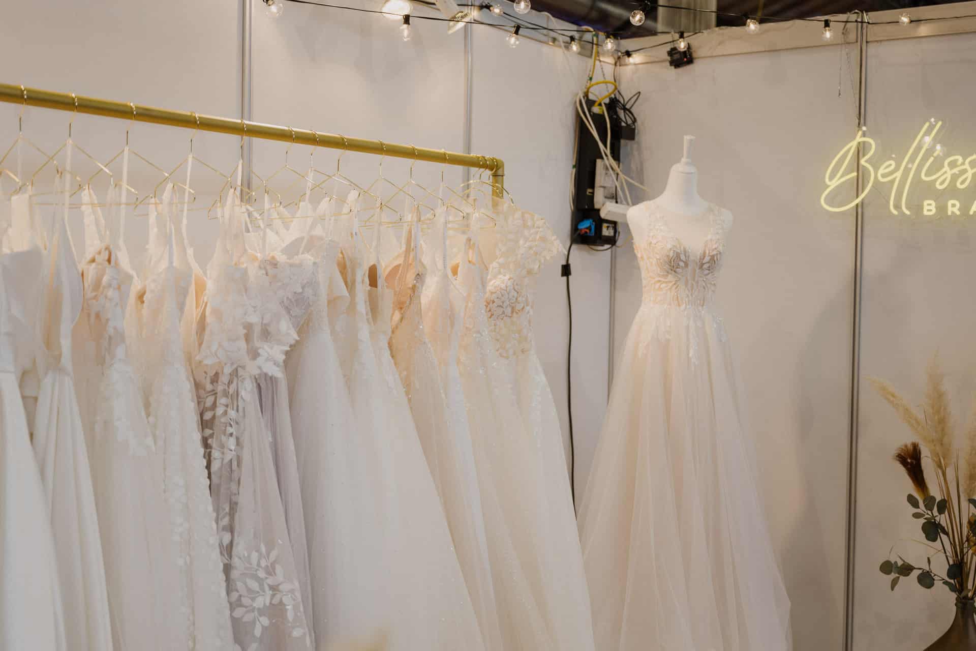 Eine Ausstellung von Brautkleidern mit verschiedenen Spitzen- und Blumenmustern auf einem Ständer verströmt den Charme von Hochzeiten. Eine Schaufensterpuppe präsentiert ein Kleid neben einem Neonschild mit der Aufschrift „Belisse“. Darüber hängen Lichterketten und beleuchten die elegante Szene für Feste.