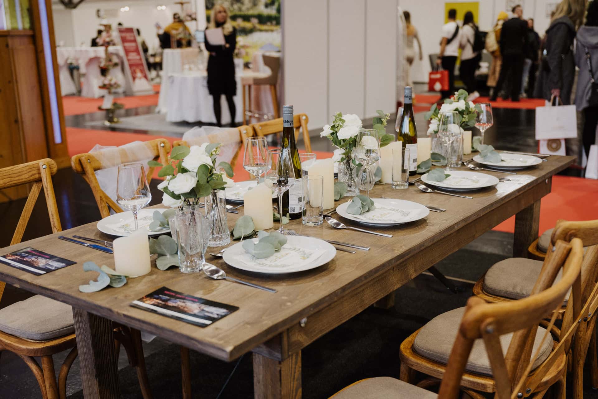 Ein Holztisch ist elegant für einen festlichen Anlass gedeckt, komplett mit weißen Tellern, Gläsern, Kerzen und Vasen mit weißen Blumen und Grünpflanzen. Weinflaschen sind kunstvoll zwischen den Gedecken verteilt. Im Hintergrund mischen sich die Leute, was an eine schicke Ausstellung oder vielleicht eine Hochzeitsfeier erinnert.