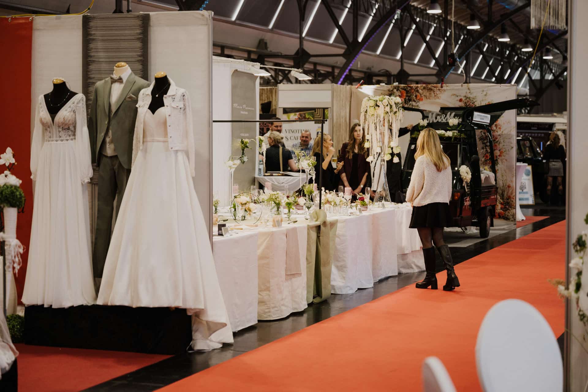 Auf einer Hochzeitsmesse herrscht geschäftiges Treiben. Schaufensterpuppen in Brautkleidung wie weißen Kleidern und hellgrauen Anzügen stehen stolz da. Eine Frau schreitet über den roten Teppich zwischen dekorierten Ständen und bestaunt atemberaubende Dekorationen, Blumenarrangements und andere Köstlichkeiten für die Braut.